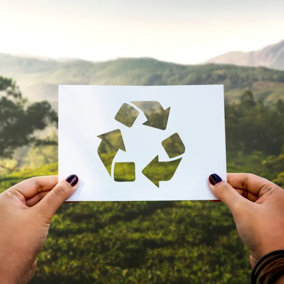 A close-up of two hands holding a Recycling sign in front of a landscape that includes pastureland and mountains.